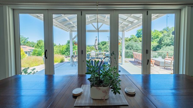 A room with a large wooden table, a potted plant, and a view through glass doors to an outdoor patio area with furniture. - Designer Ranch w/Gourmet Kitchen and Downtown Views. (Paso Robles)