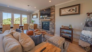 A living room with a stone fireplace, a large flat-screen TV mounted above it, and a cozy seating area with a sofa and ottoman. - Designer Ranch w/Gourmet Kitchen and Downtown Views. (Paso Robles)