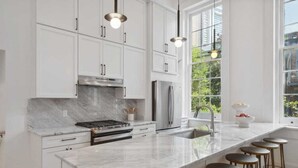 A modern kitchen with white cabinets, a marble countertop, a stainless steel range, and a large window providing a view of greenery outside.
