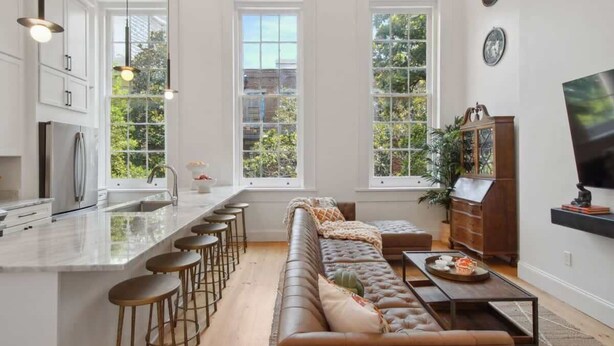 A modern kitchen with a central island, bar stools, a long brown sofa, and a dining table with a tray of food.