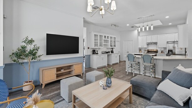 A modern living room with a flat-screen TV, a dining area, and a kitchen with white cabinets.