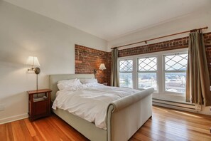 A bedroom with a bed, a nightstand, a lamp, a window with curtains, and a brick wall. - Modern 2BR Loft: Walk to Downtown - 30 plus day (Leadville)