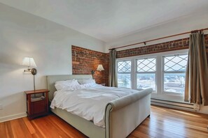 A bedroom with a bed, a nightstand, a wall-mounted lamp, a brick accent wall, and a large window with curtains. - Modern 2BR Loft: Walk to Downtown - 30 plus day (Leadville)