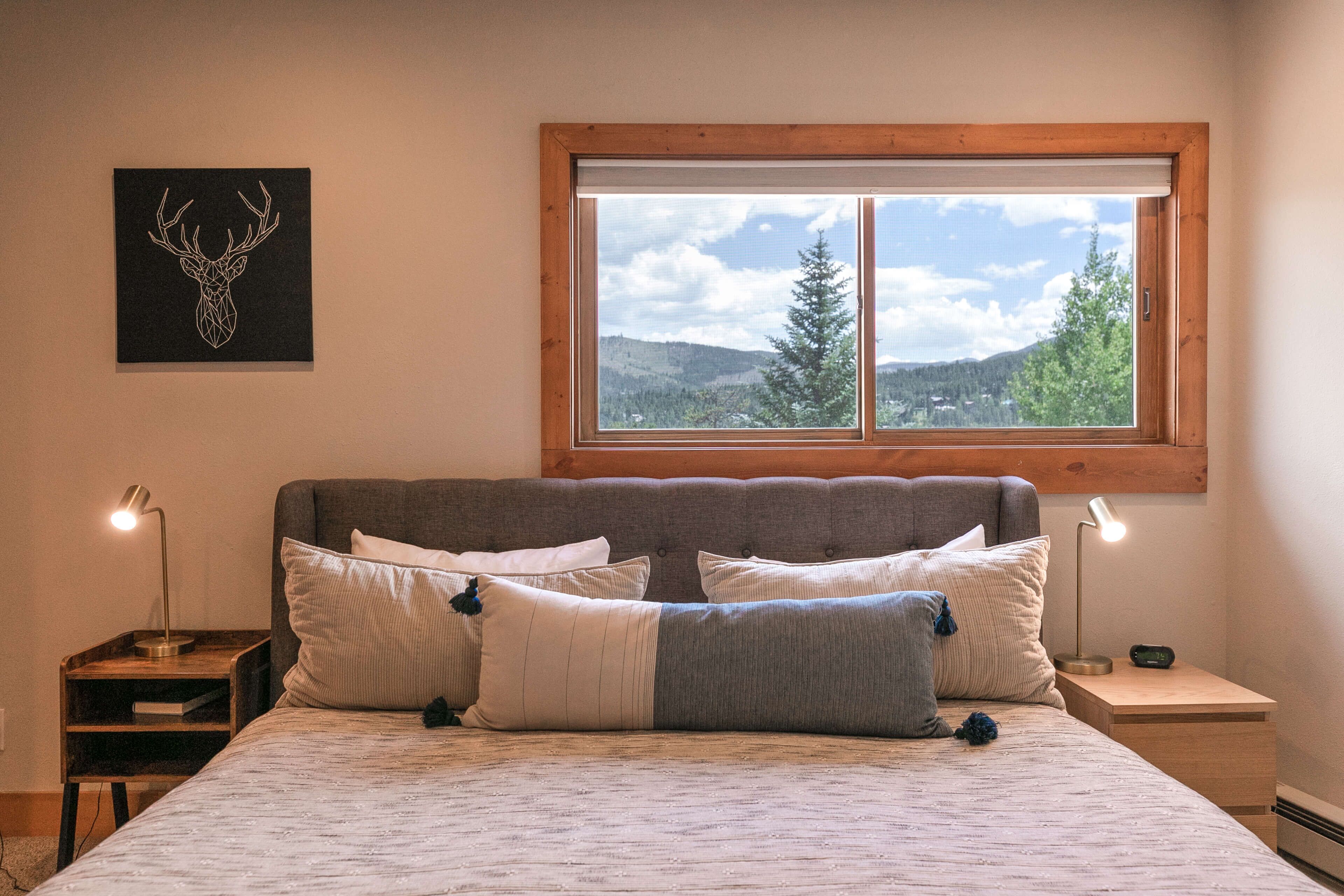 A bedroom with a bed, pillows, a nightstand, a lamp, and a window with a view of trees and mountains.