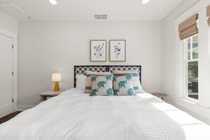 Una cama bien hecha con ropa de cama blanca, dos cuadros enmarcados, una lámpara de noche y una ventana con persianas.