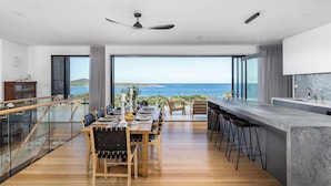 A modern kitchen with a large island, dining area with a view of the sea, and a staircase leading to an upper level. - Fingal Bay Panorama - Panoramic Ocean Views, 300m to Beach with Pool Table (Fingal Bay)