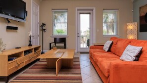 A living room with a TV, wooden entertainment unit, orange sofa, and striped rug. - Sleeps 6 in a 4plex w Pool, Only a Block to Beach (South Padre Island)