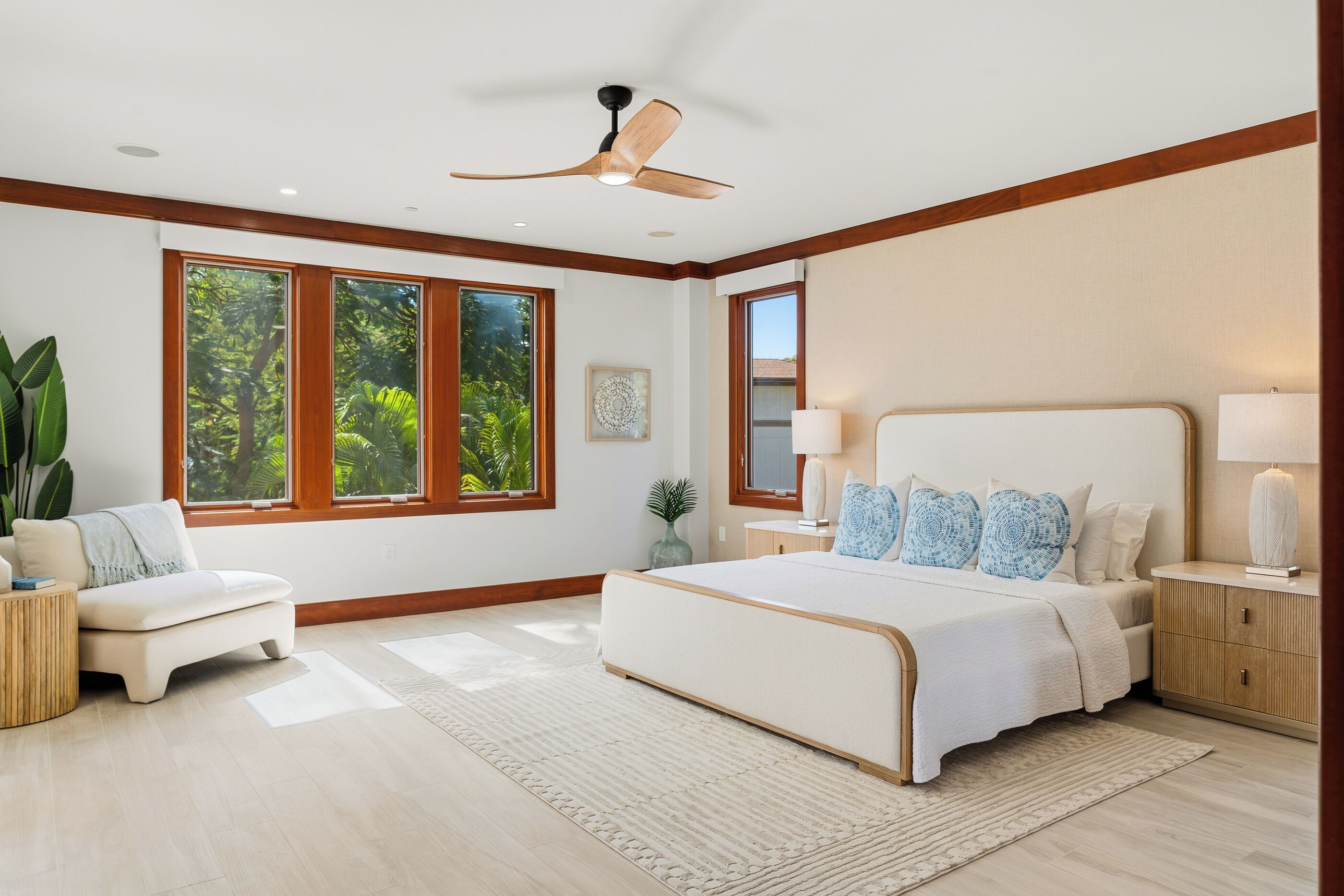 A bedroom with a large bed, a ceiling fan, and a view of greenery through the windows.