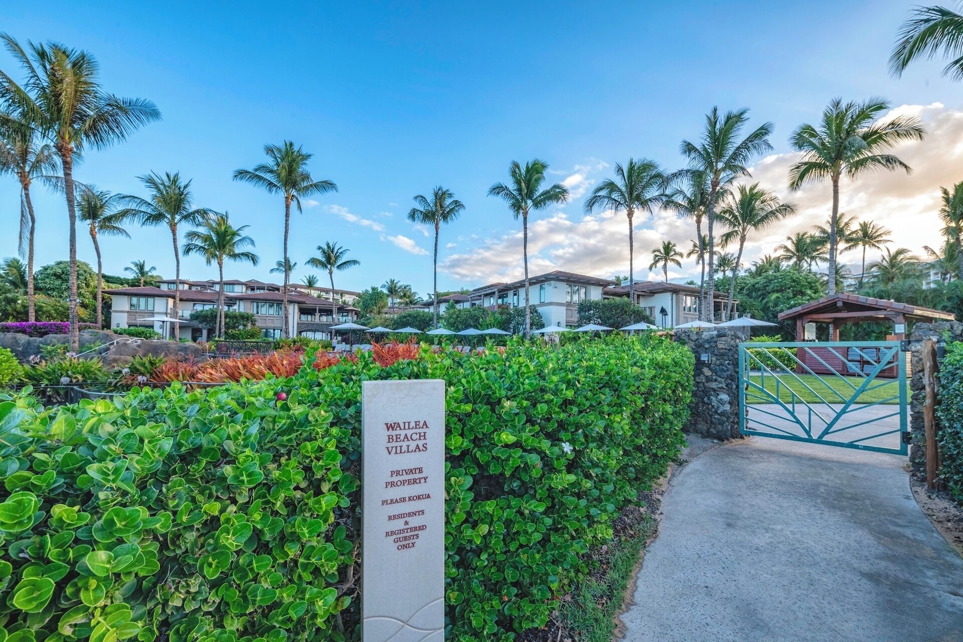 A private property entrance with a sign indicating 'Wailele Beach Villas' and a gated area with lush greenery and palm trees.