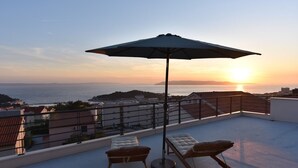 Una terrazza panoramica con due poltrone da relax e un ombrellone, con vista su una città costiera al tramonto.