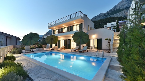 Una moderna casa a due piani con piscina, area salotto esterna e montagne sullo sfondo.