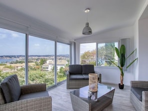 Uma sala de estar moderna com janelas amplas, um sofá preto, móveis de vime e um lustre.
