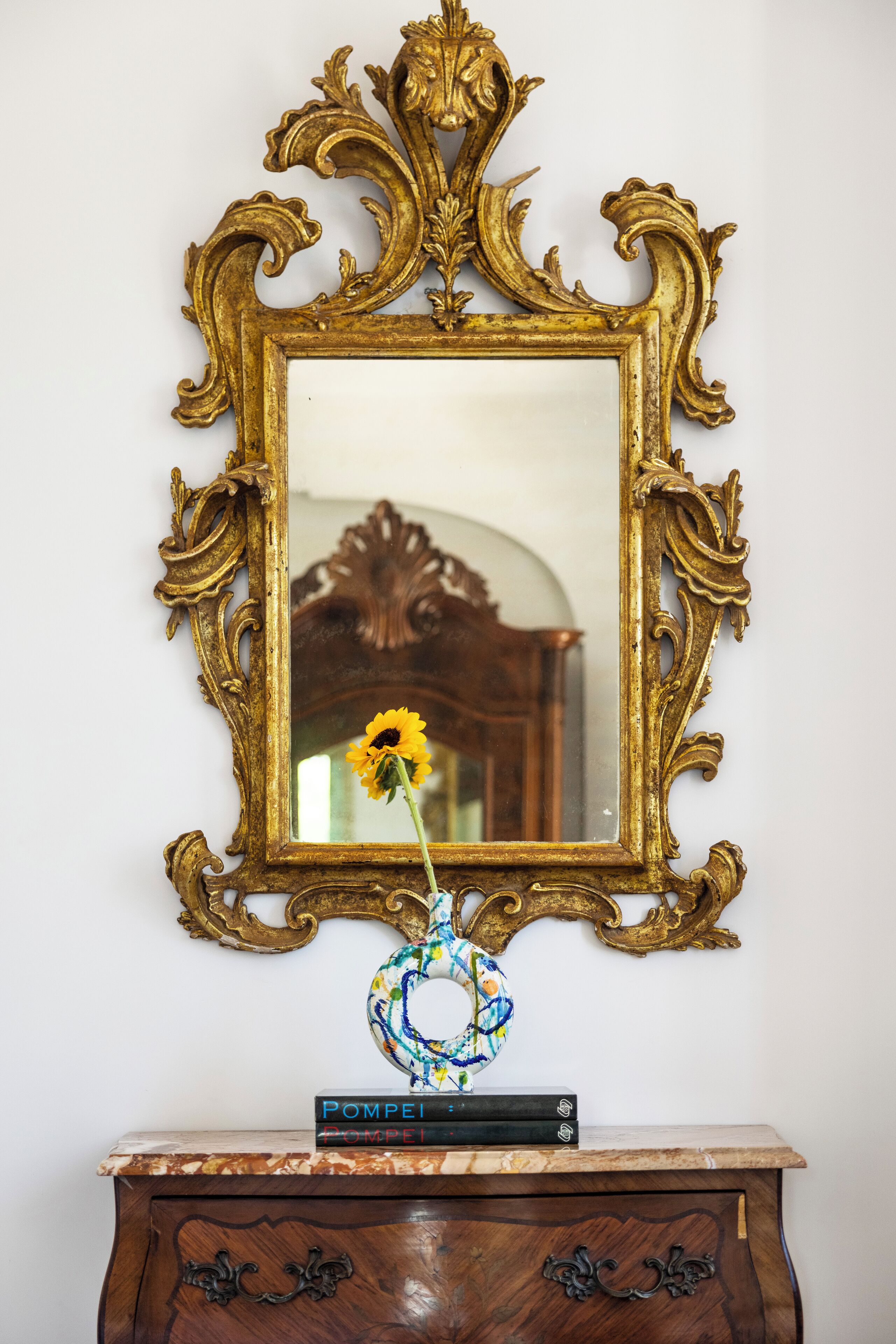 A mirror with an ornate gold frame, a vase with a sunflower, and a stack of books on a wooden dresser.