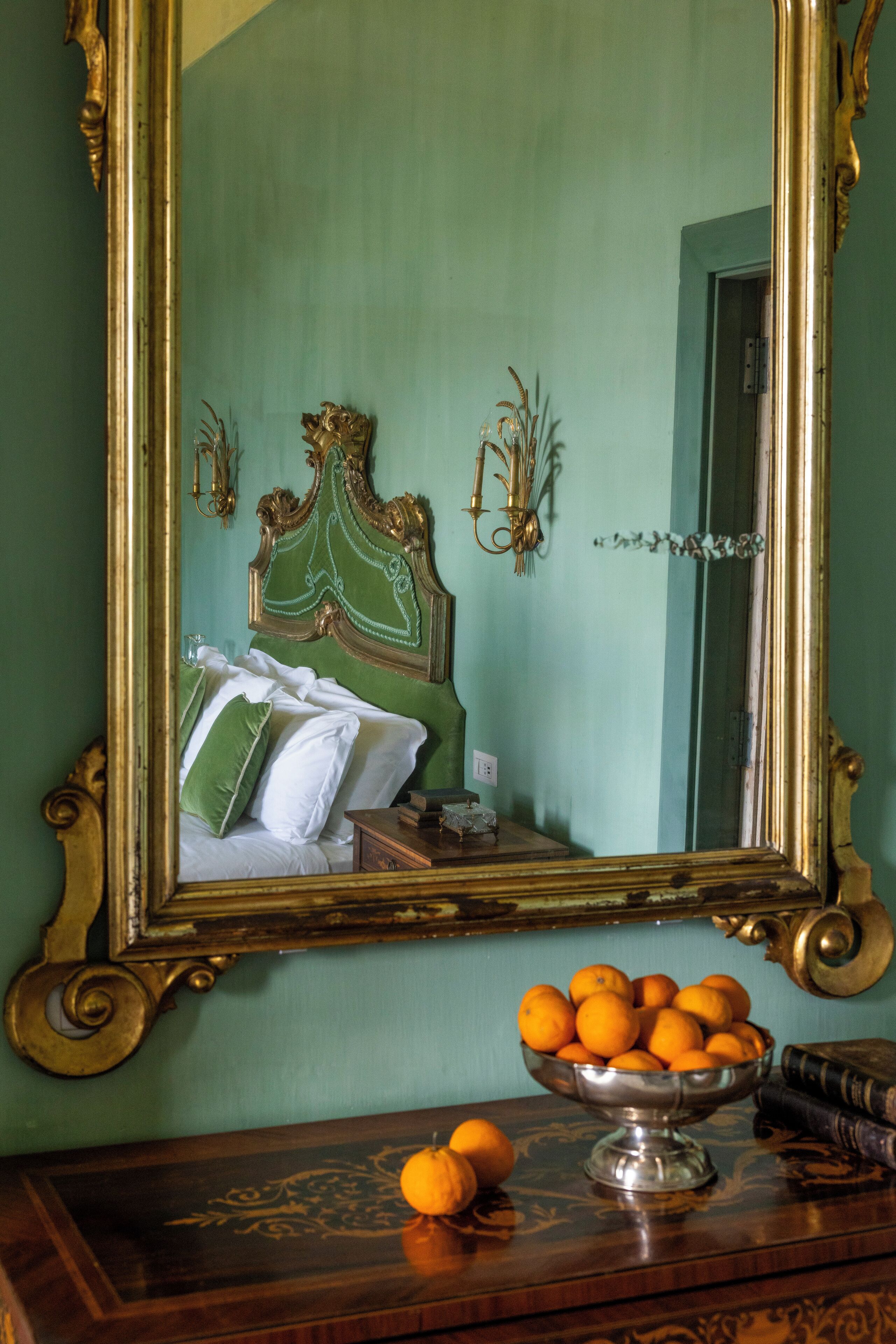 A room with a green wall, a bed with white and green pillows, a wooden dresser, a mirror with an ornate frame, and a bowl of oranges on the dresser.