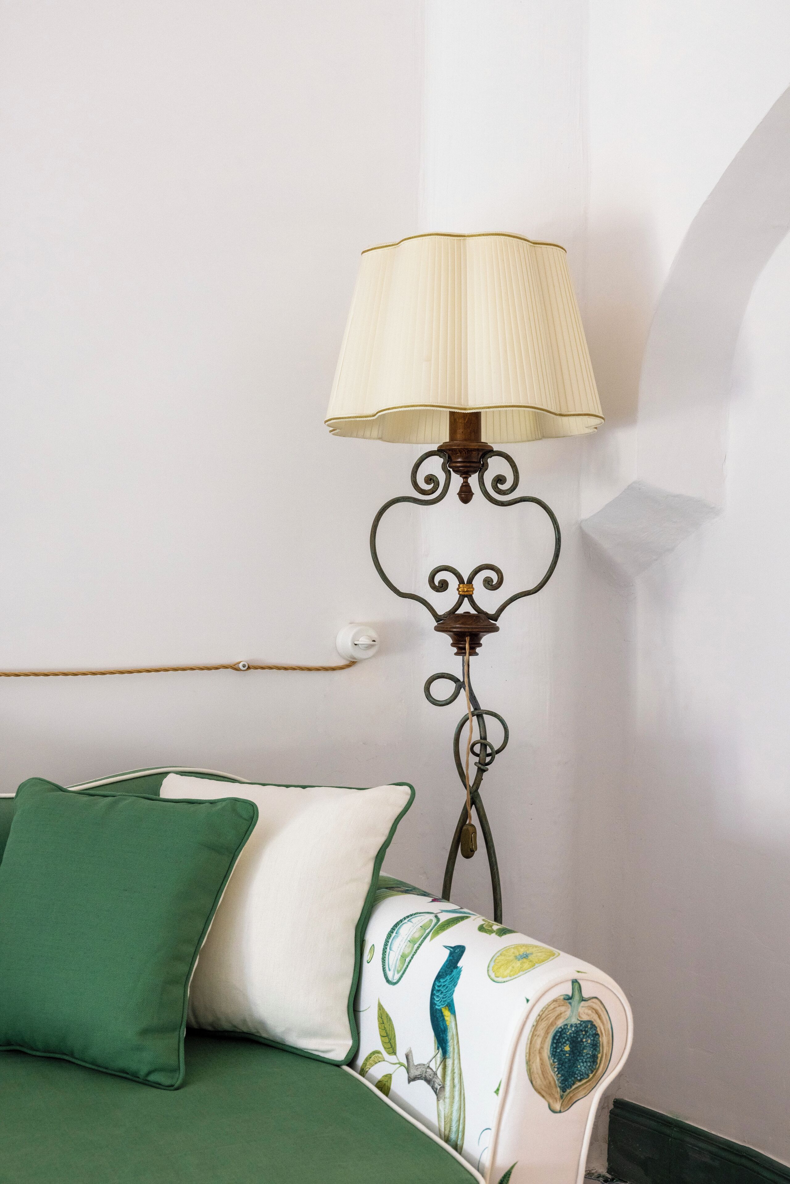 Um sofá verde com uma almofada branca e uma almofada estampada, uma luminária na parede com cúpula bege, e uma parede branca com uma pequena janela.
