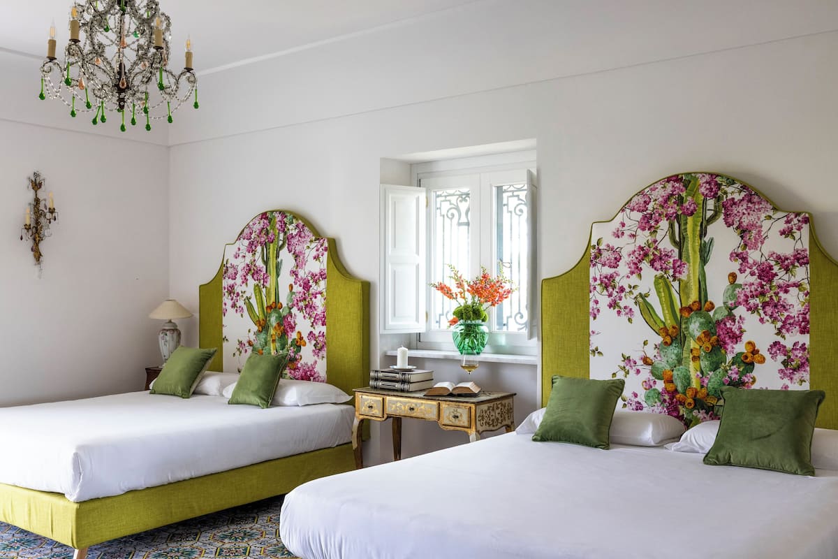 Habitación de hotel con dos camas, un candelabro, una ventana con vistas y una mesita con libros y un florero.