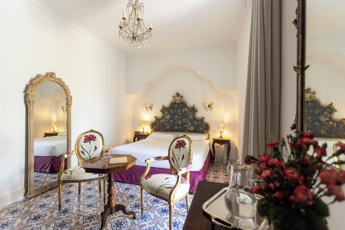 Habitación de hotel con cama, dos sillas, una mesa, candelabro y espejo.