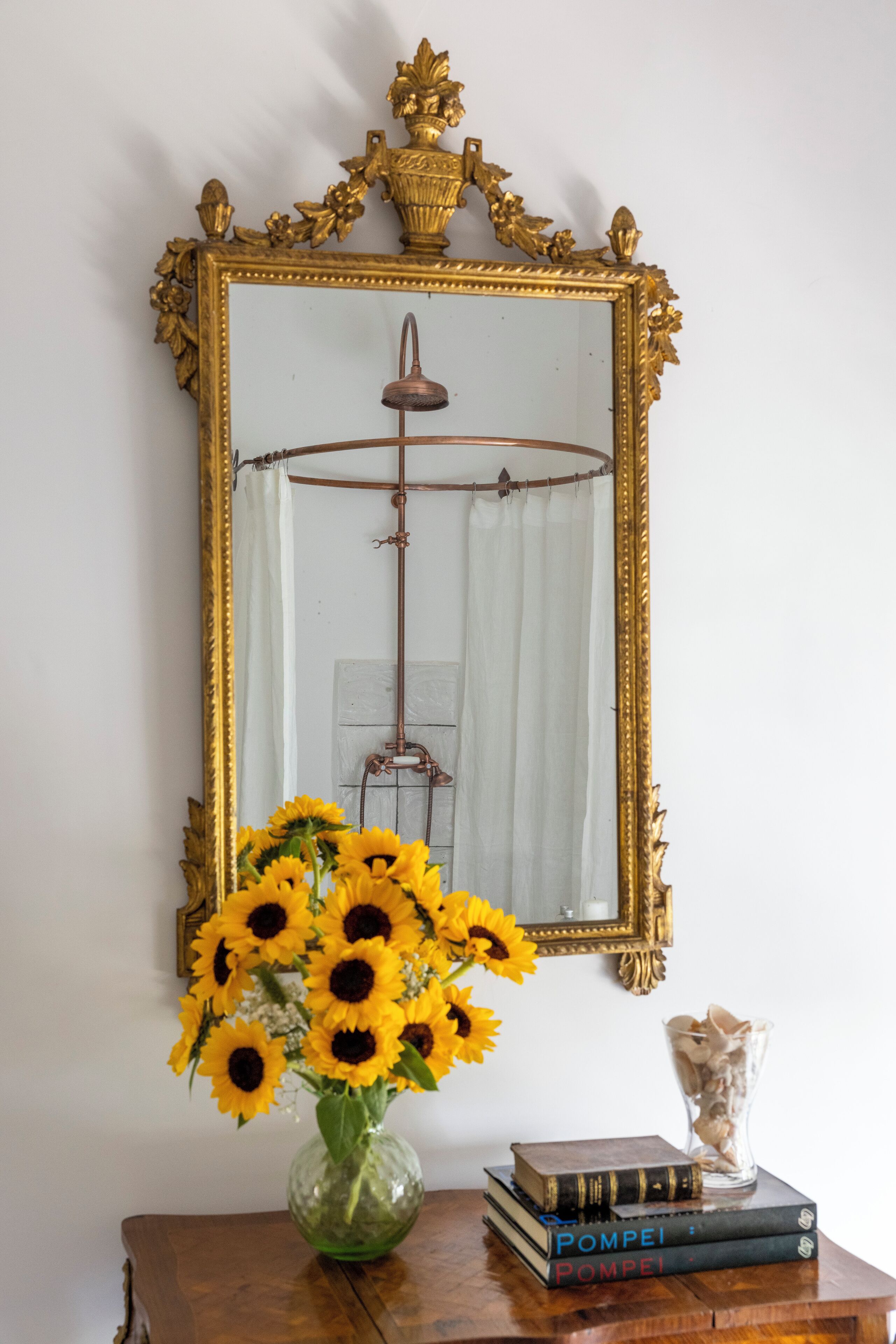 Ein Spiegel mit goldener Rahmung, eine Vase mit Sonnenblumen und ein Stapel Bücher auf einem Holztisch.