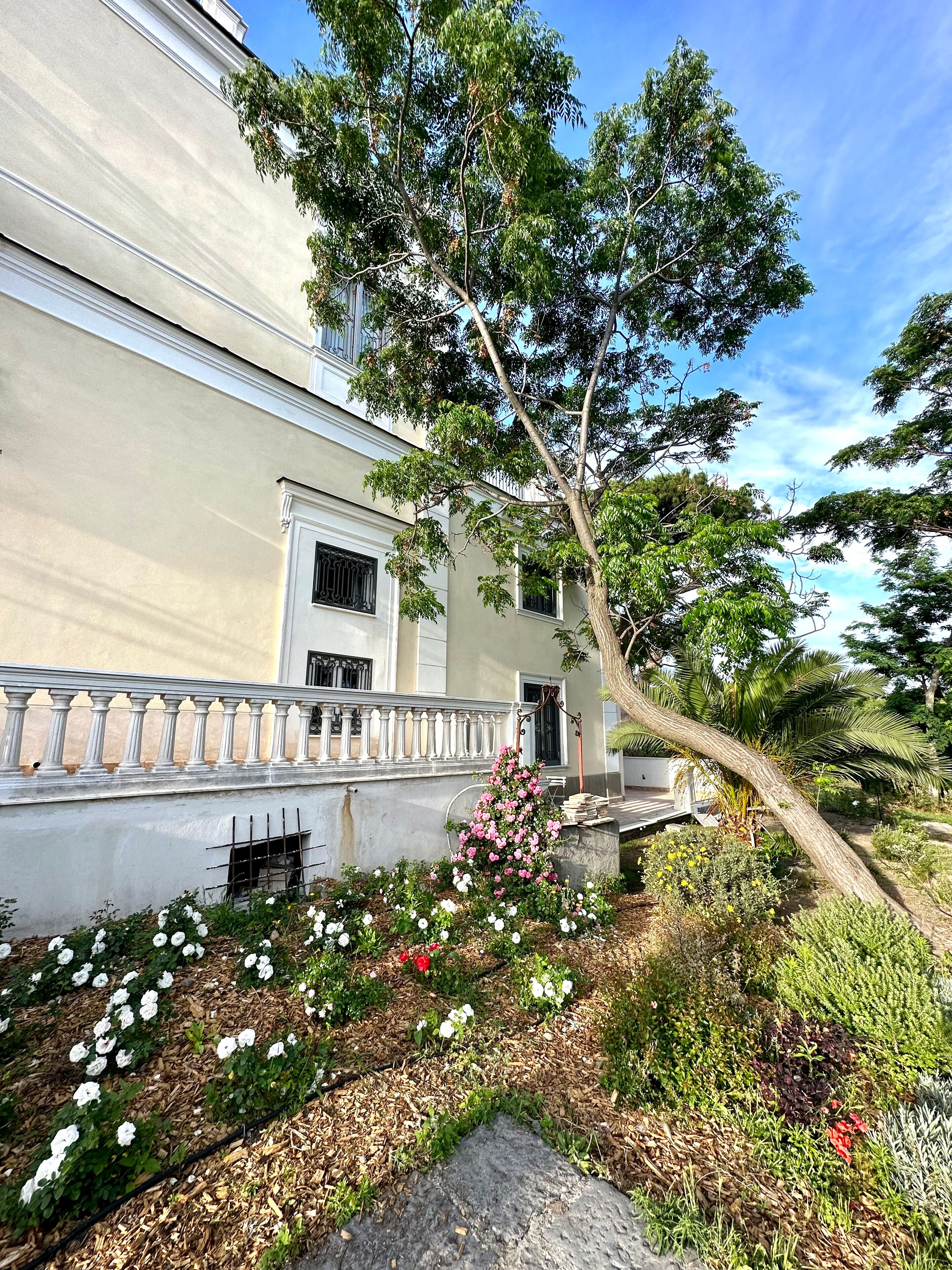 A building with a white facade, a balcony with balustrades, and a garden with various plants and flowers.