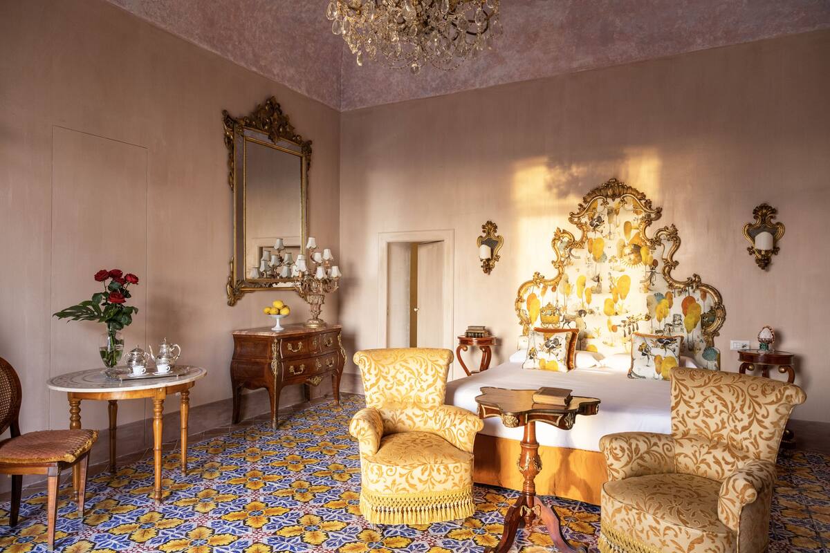 Una habitación con una cama, dos sillones, una mesa, un espejo y una araña.