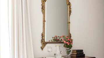 Une pièce comprenant un grand miroir décoratif, une commode en bois, une table ronde avec deux verres et une chaise recouverte d’un tissu à motifs floraux.