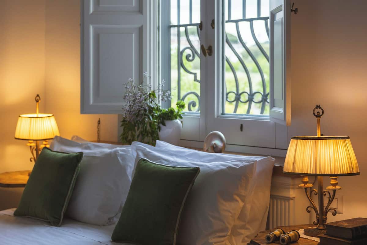Una cama bien hecha con almohadas verdes, dos lámparas de noche y una ventana con vistas a la vegetación.