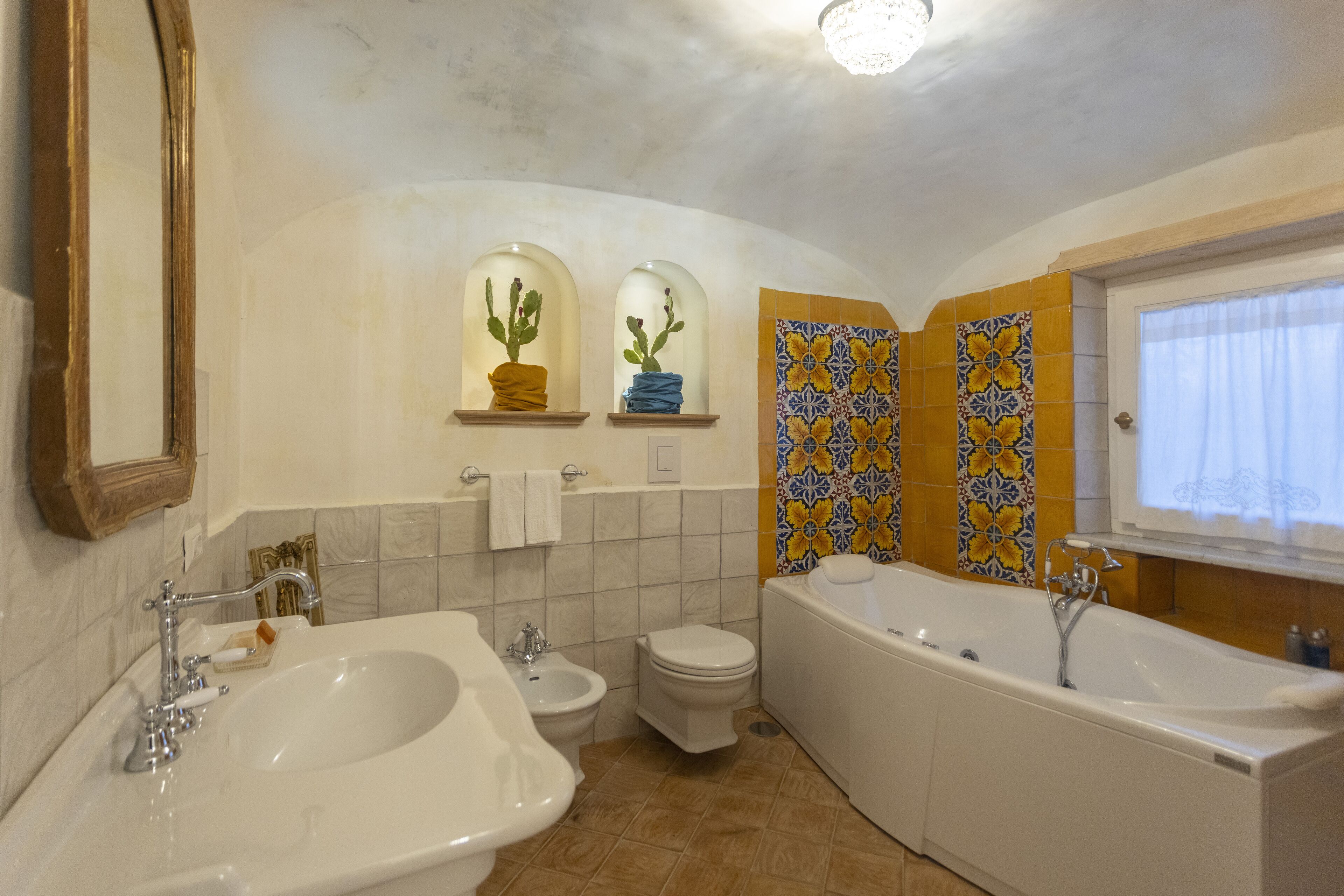 Un baño con bañera, dos lavamanos, inodoro y bidé. Hay plantas decorativas en nichos de ventana y azulejos estampados en las paredes.