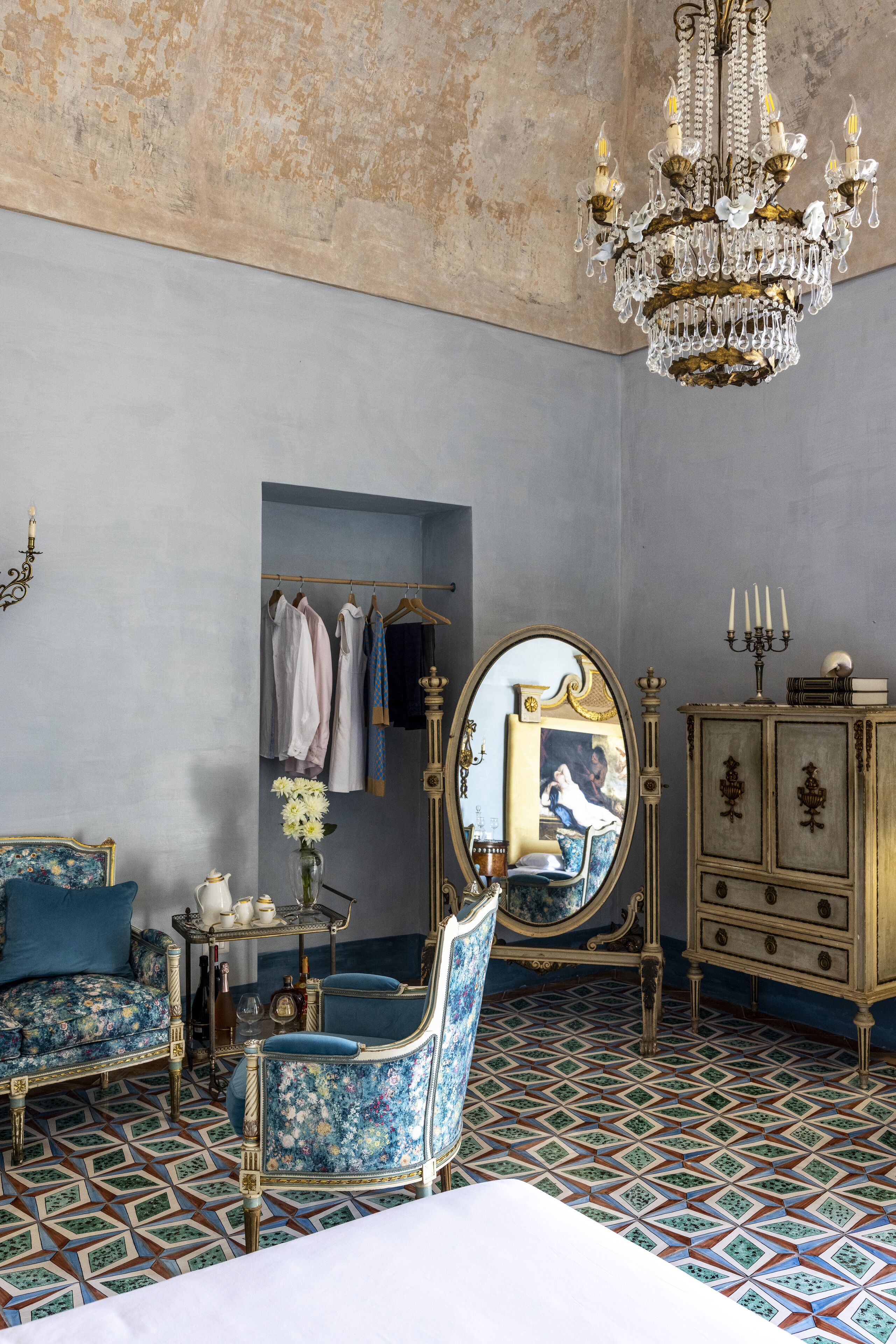 A room with a bed, a blue armchair, a dresser, a mirror, and a chandelier.