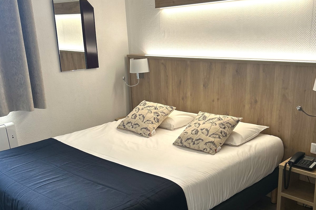 A neatly made bed with a dark blue bedspread, two patterned pillows, and a wooden headboard. A bedside table with a lamp and a phone is visible to the right.