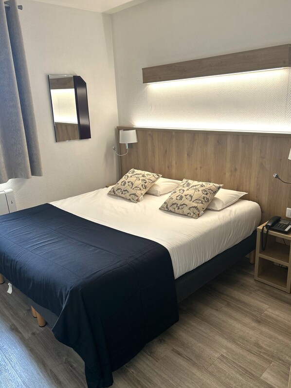A neatly made bed with a dark blue bedspread, two patterned pillows, and a wooden headboard. A bedside table with a lamp and a phone is visible to the right.