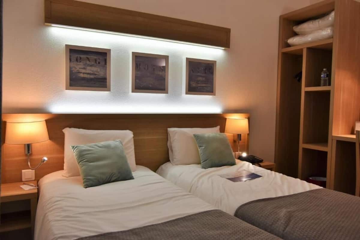 A hotel room with two beds, each with white bedding and green pillows. There are wall-mounted lamps and a wooden headboard.