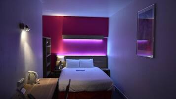 Ein Hotelzimmer mit Bett, Nachttisch, Lampe und einem an der Wand befestigten Kunstwerk.