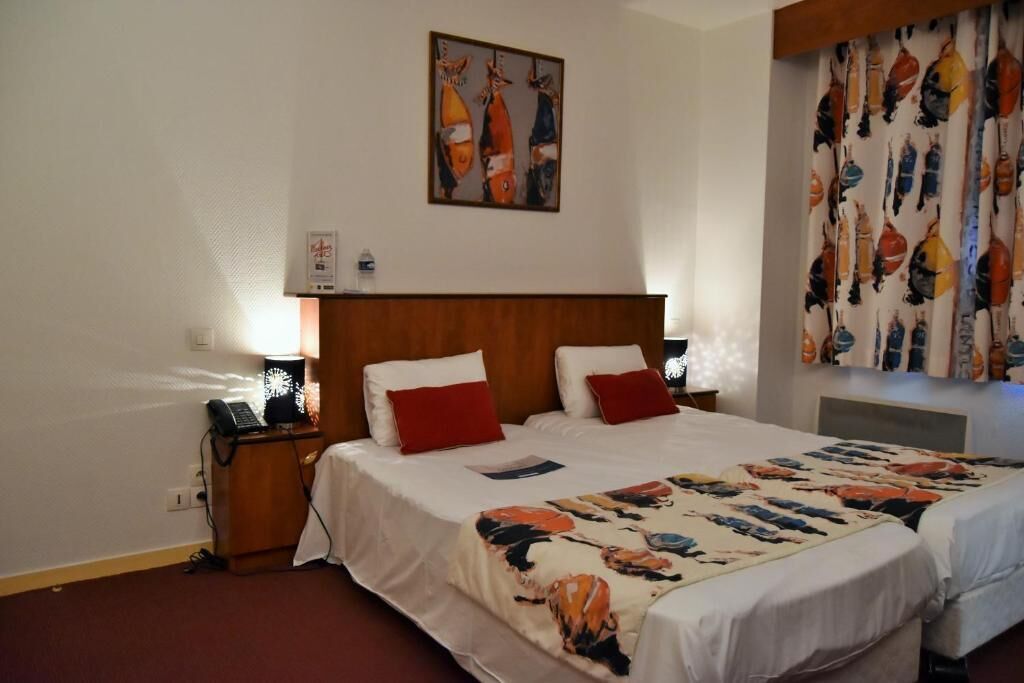 Quarto de hotel com cabeceira de madeira, duas camas com roupa de cama branca e vermelha, um quadro abstrato colorido na parede e cortina estampada.