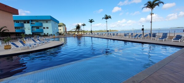 A swimming pool with lounge chairs and palm trees, surrounded by buildings with balconies. - Golden entre o mar e a lagoa (Arraial do Cabo)