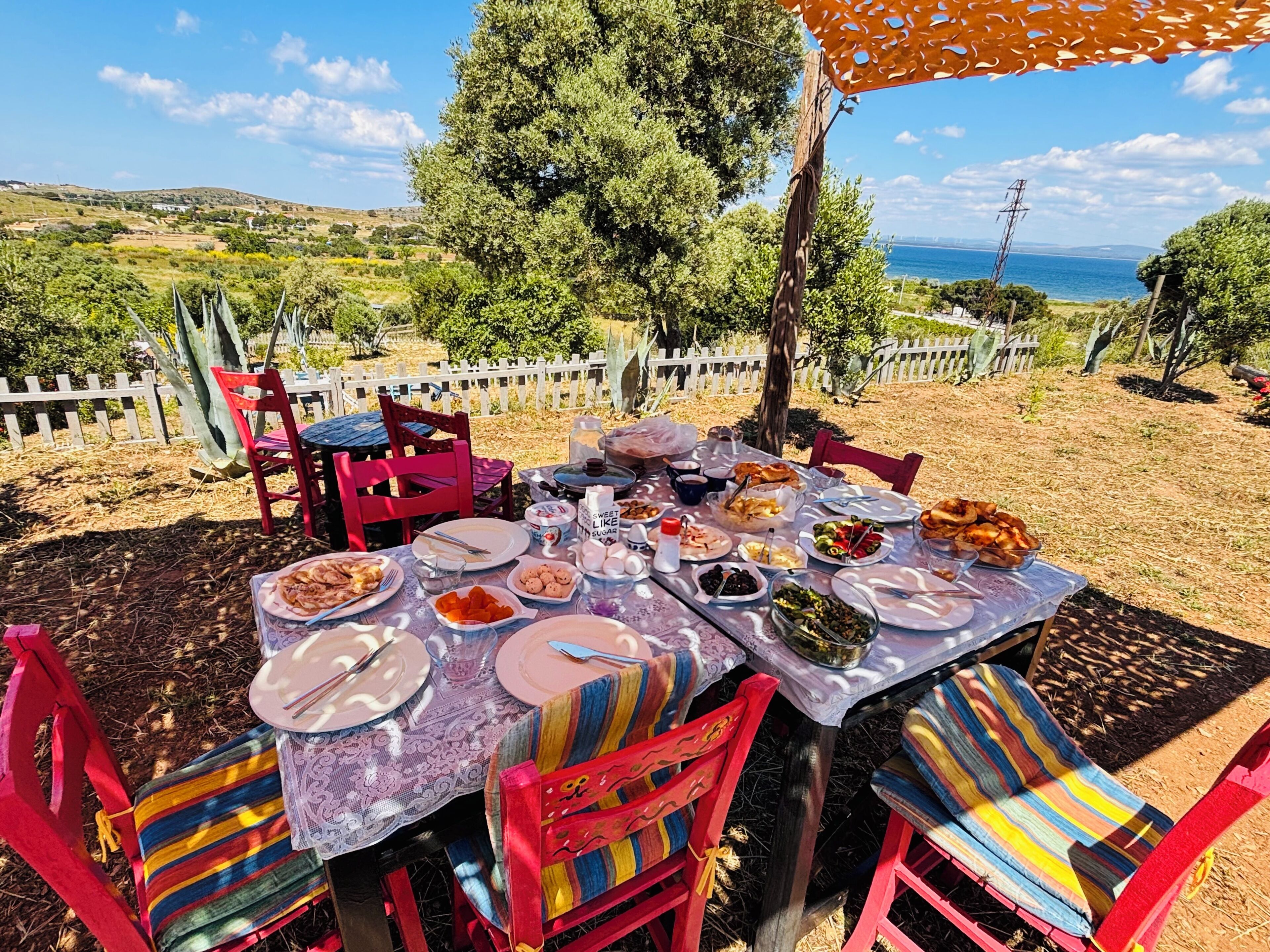 An outdoor dining area with a table set for a meal, surrounded by chairs, and a view of the sea and hills in the background.