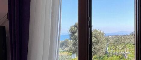 Une pièce dotée d’une grande fenêtre, de rideaux et offrant une vue sur un paysage verdoyant, avec des arbres et une étendue d’eau.