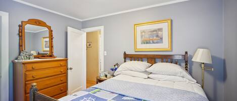 A bedroom with a wooden bed, a dresser, a nightstand, a framed picture on the wall, and a lamp.
