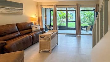A living room with a brown leather sofa, a wicker coffee table, and a view of a garden through large glass doors.