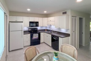 A modern kitchen with white cabinets, a black stove, a microwave, and a dishwasher. There is a glass table with chairs and a decorative pineapple centerpiece. - Cozy Condo w/ Pool Access 1 Mi From Siesta Key (Sarasota)