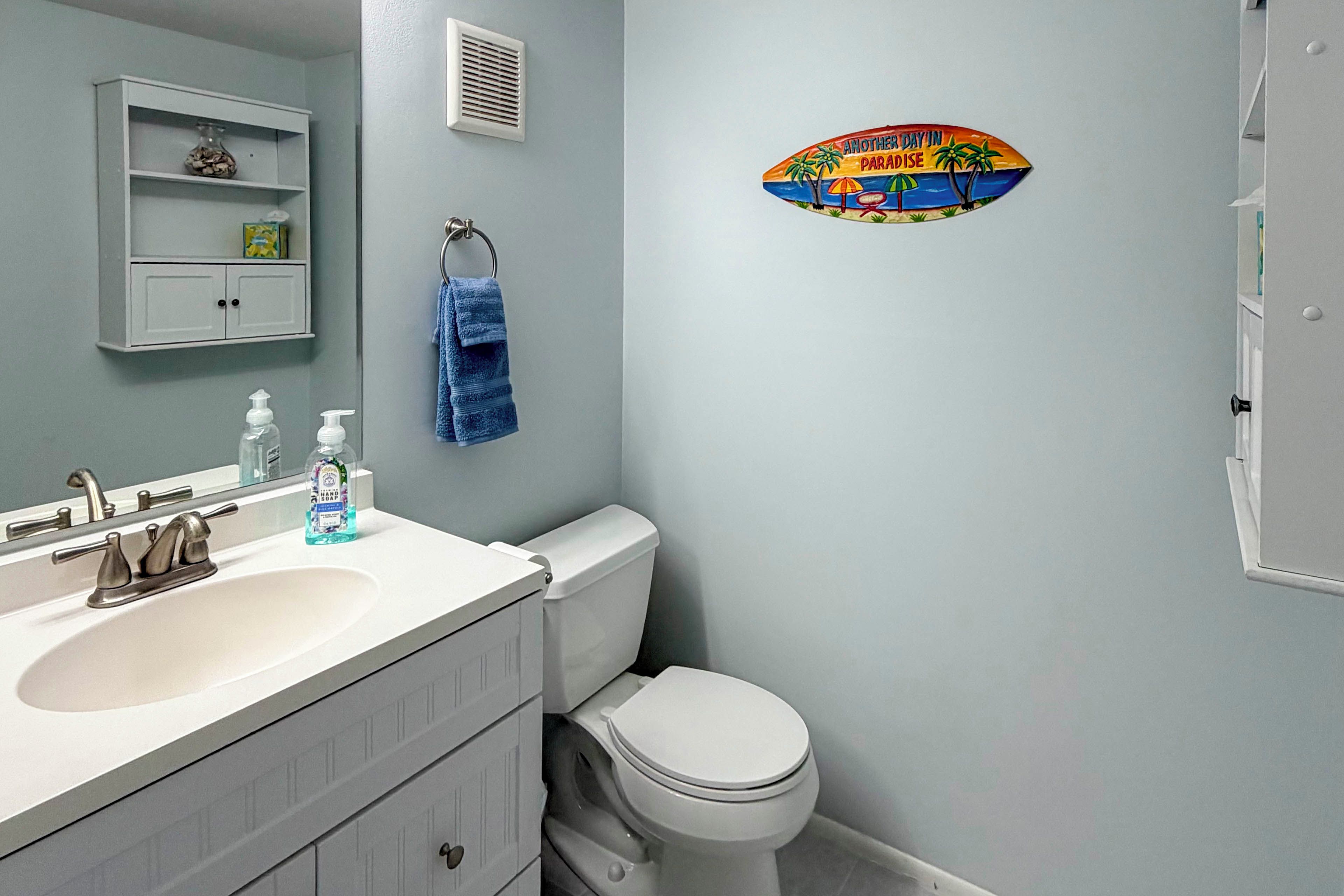 Een badkamer met een witte wastafel, een toilet, een handdoek aan een ring, een zeepdispenser en een vent op de muur.