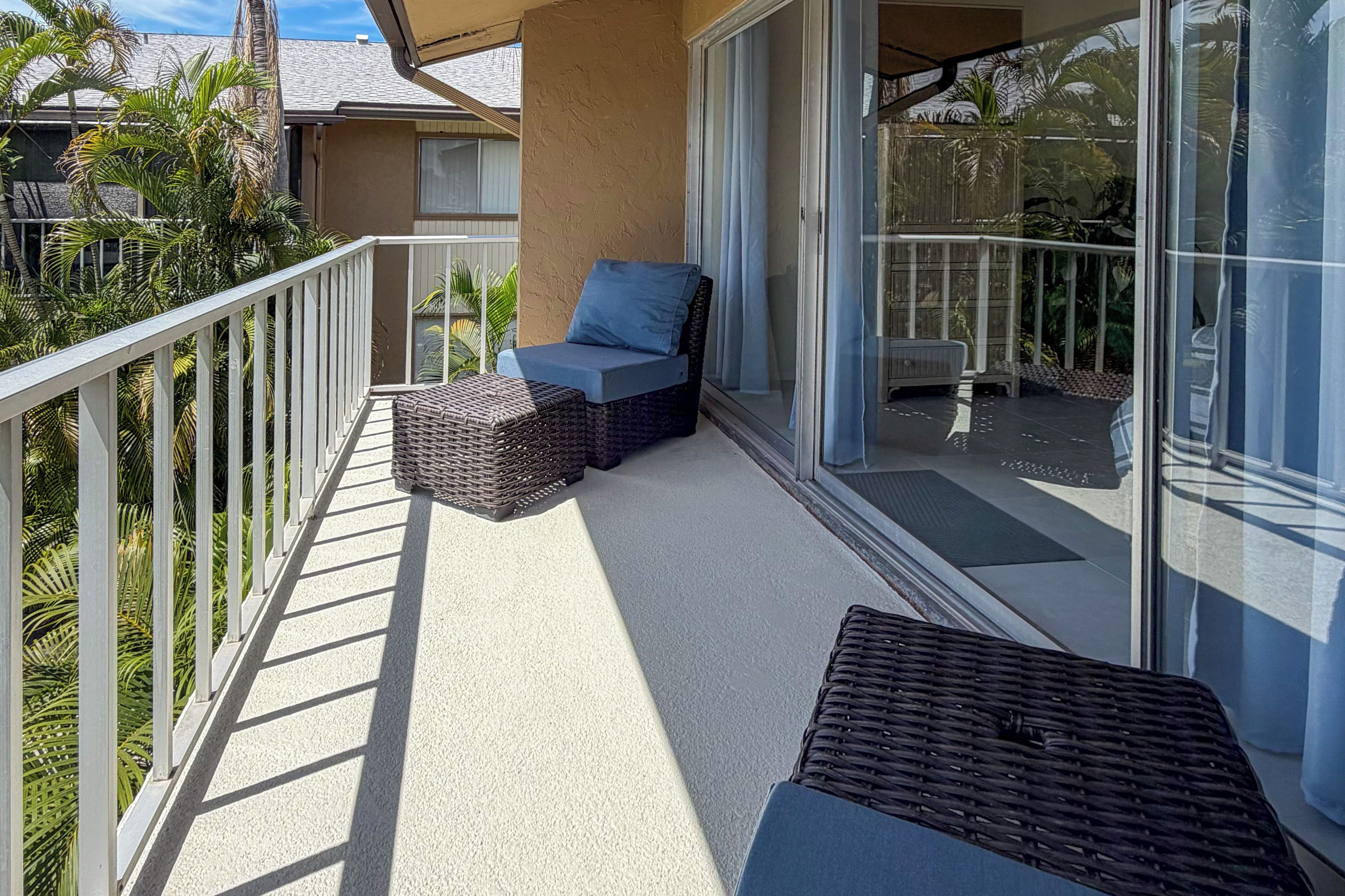 A balcony with a wicker sofa and ottoman, a glass door, and a view of a house and palm trees.