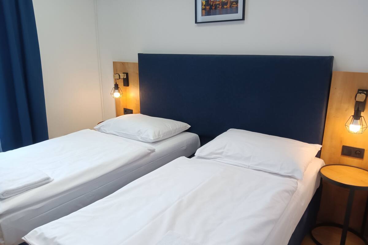Two single beds with white linens, a dark blue headboard, and a small round table with a lamp.