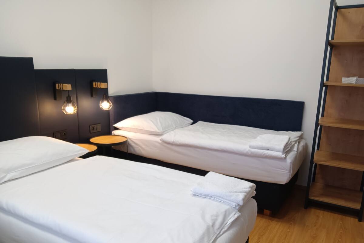 A hotel room with two single beds, a small wooden table, and a nightstand with a lamp.