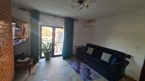 A living room with a dark blue sofa, a potted plant, a patterned rug, and a wall-mounted TV displaying a show.