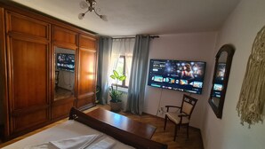 A room with a wooden wardrobe, a bed, a television mounted on the wall, a potted plant, and a chair.