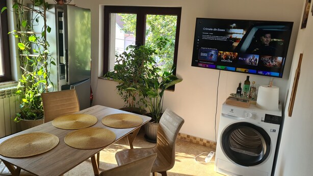 A dining area with a table set for two, a washing machine, and a TV mounted on the wall displaying a movie.