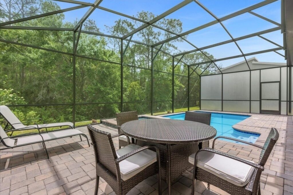 Un patio exterior con piscina, rodeado por un recinto de vidrio, con mobiliario de mimbre y vistas a una zona boscosa.
