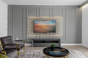 A modern living room with a large flat-screen TV mounted on a textured wall, a grey sofa, a black coffee table with a plant, and a minimalist console table. - 9 Bedroom Champions Gate Home by RedAwning (Davenport)