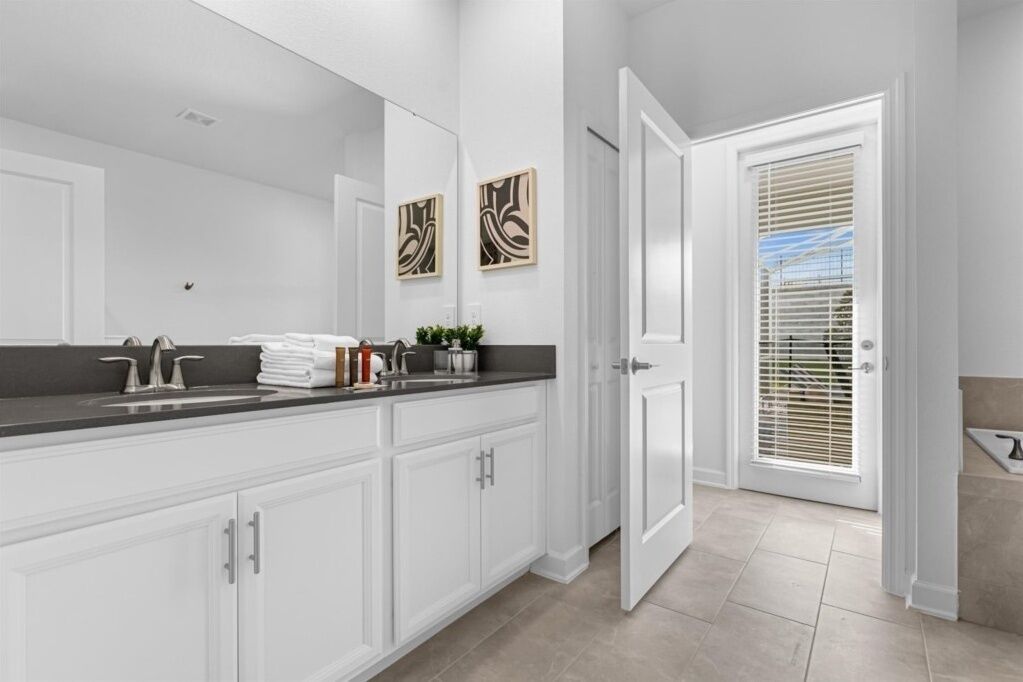 A modern bathroom with a large mirror, white cabinets, a black countertop, and a door leading to another room.