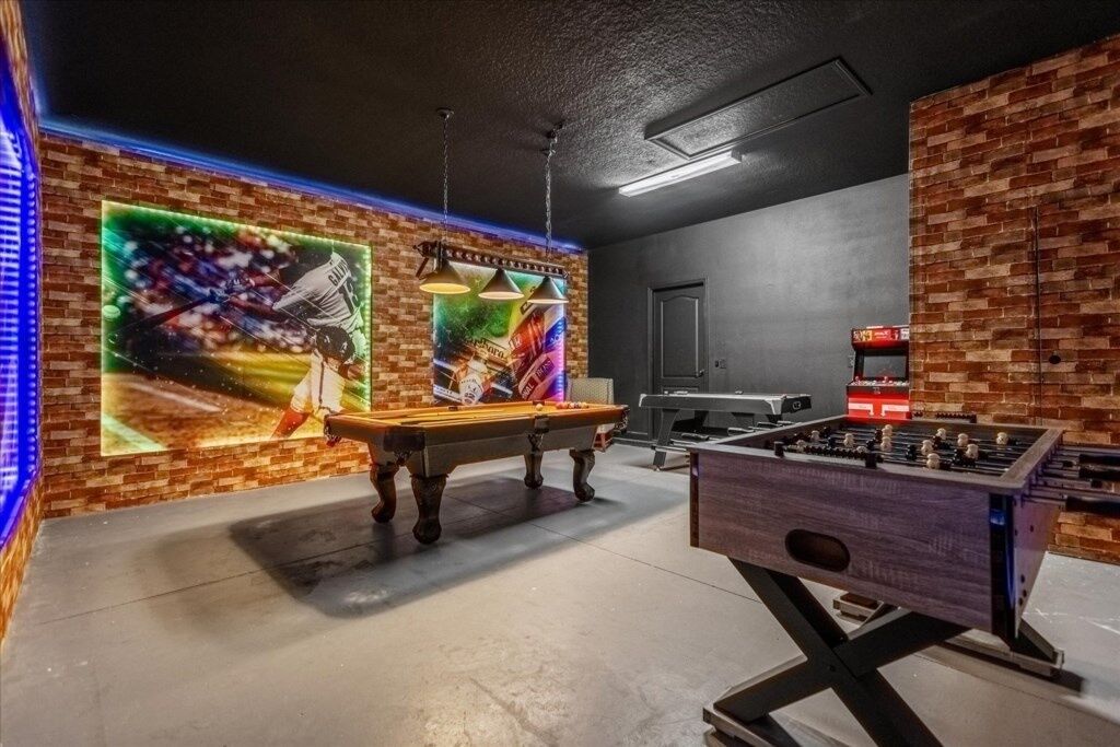 A billiards table, a foosball table, and a dartboard in a room with brick walls and a concrete floor.